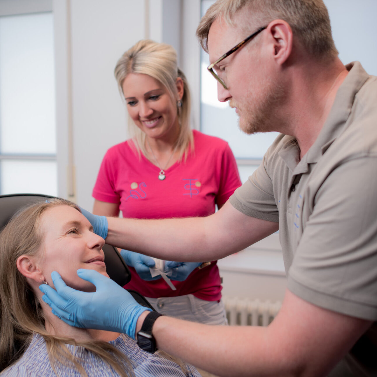 Dr. Thorsten Schmidt untersucht das Gesicht einer auf dem Stuhl liegenden Patientin während eine Angestellte ihm assistiert