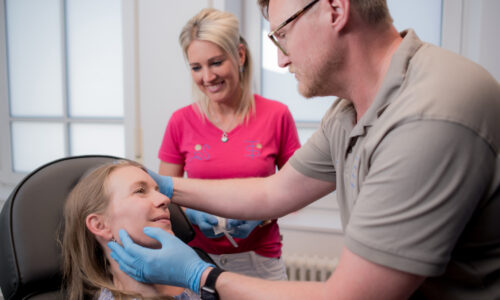 Dr. Thorsten Schmidt untersucht das Gesicht einer auf dem Stuhl liegenden Patientin während eine Angestellte ihm assistiert